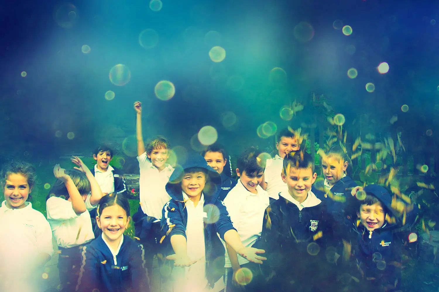 A group of children in school uniforms stand together outside, some smiling and raising their arms. The image has a colorful, out-of-focus effect with light spots.