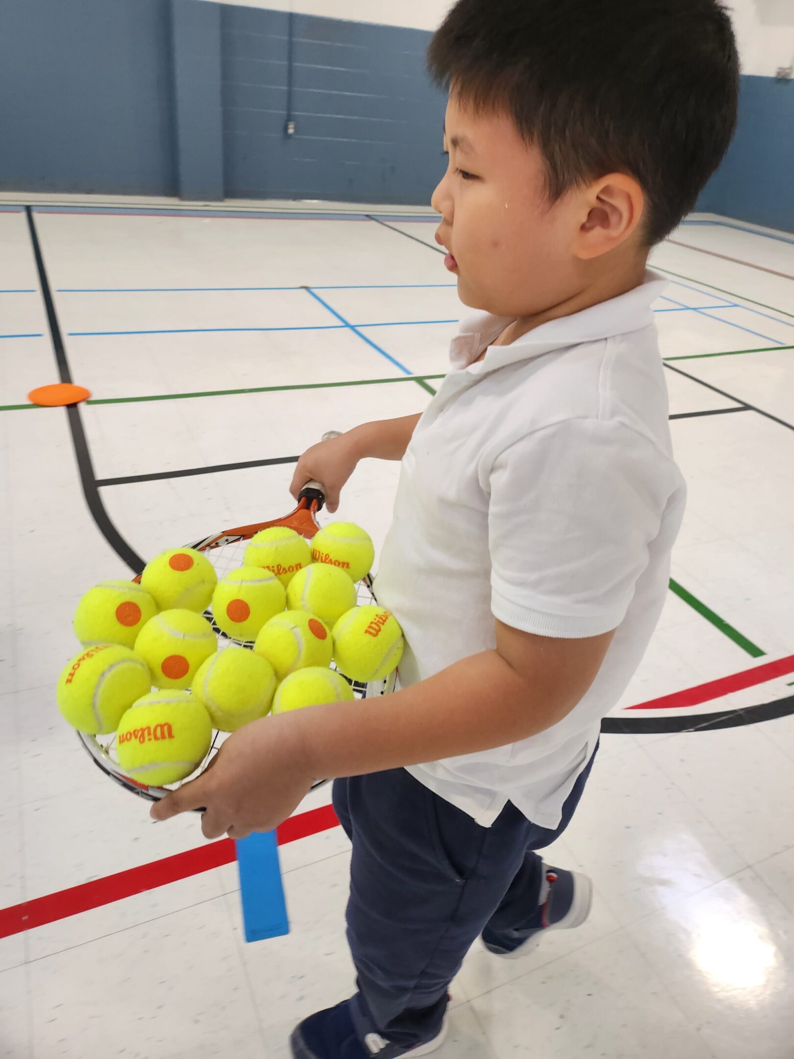 Child carrying tennis balls in gymnasium.